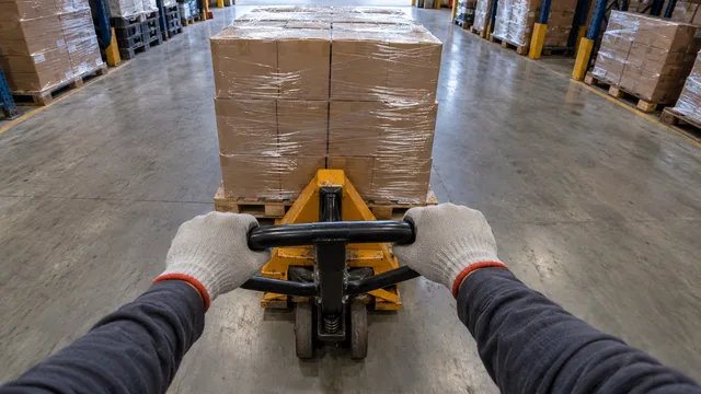 Cámara 6: dos manos empujan una transpaleta con cajas apiladas por un pasillo de almacén.