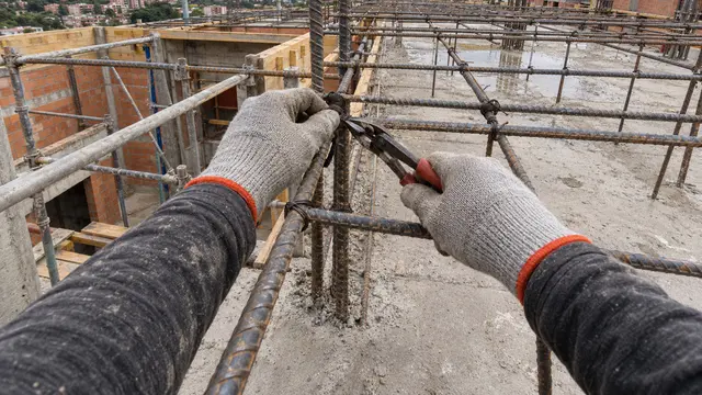 Cámara 2: dos manos con guantes grises amarran dos varillas con alambre y pinzas en una obra.